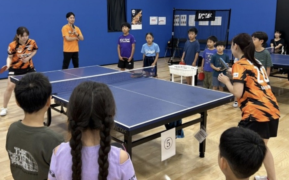 卓球台で向かい合う選手と見守る小学生たち